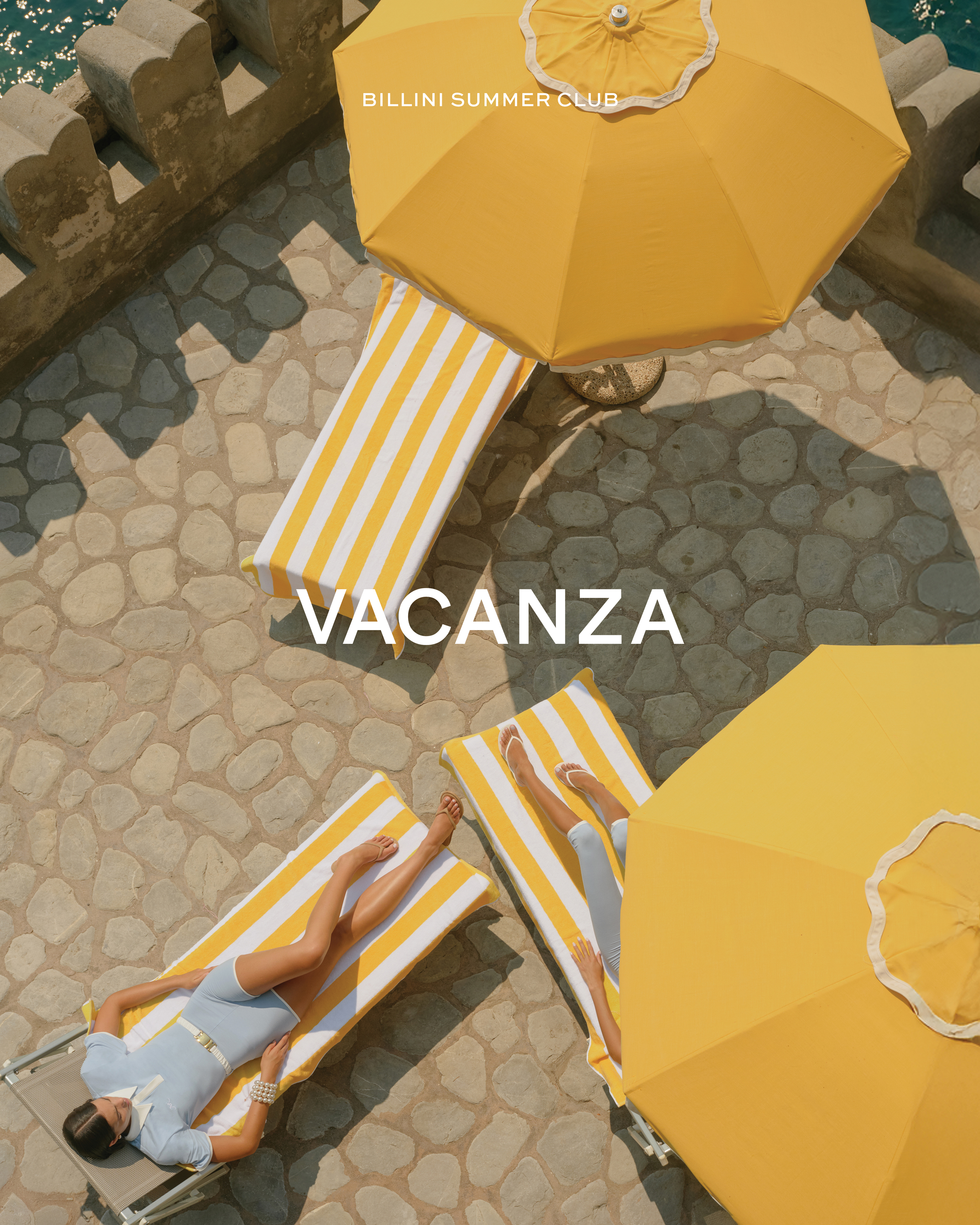 Overhead view of two women lounging on striped beach chairs under yellow umbrellas on a sunny stone patio by the sea.
