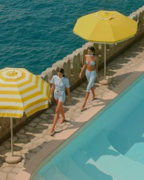 Two women walking along a stone pool deck beside blue water under yellow and white striped umbrellas