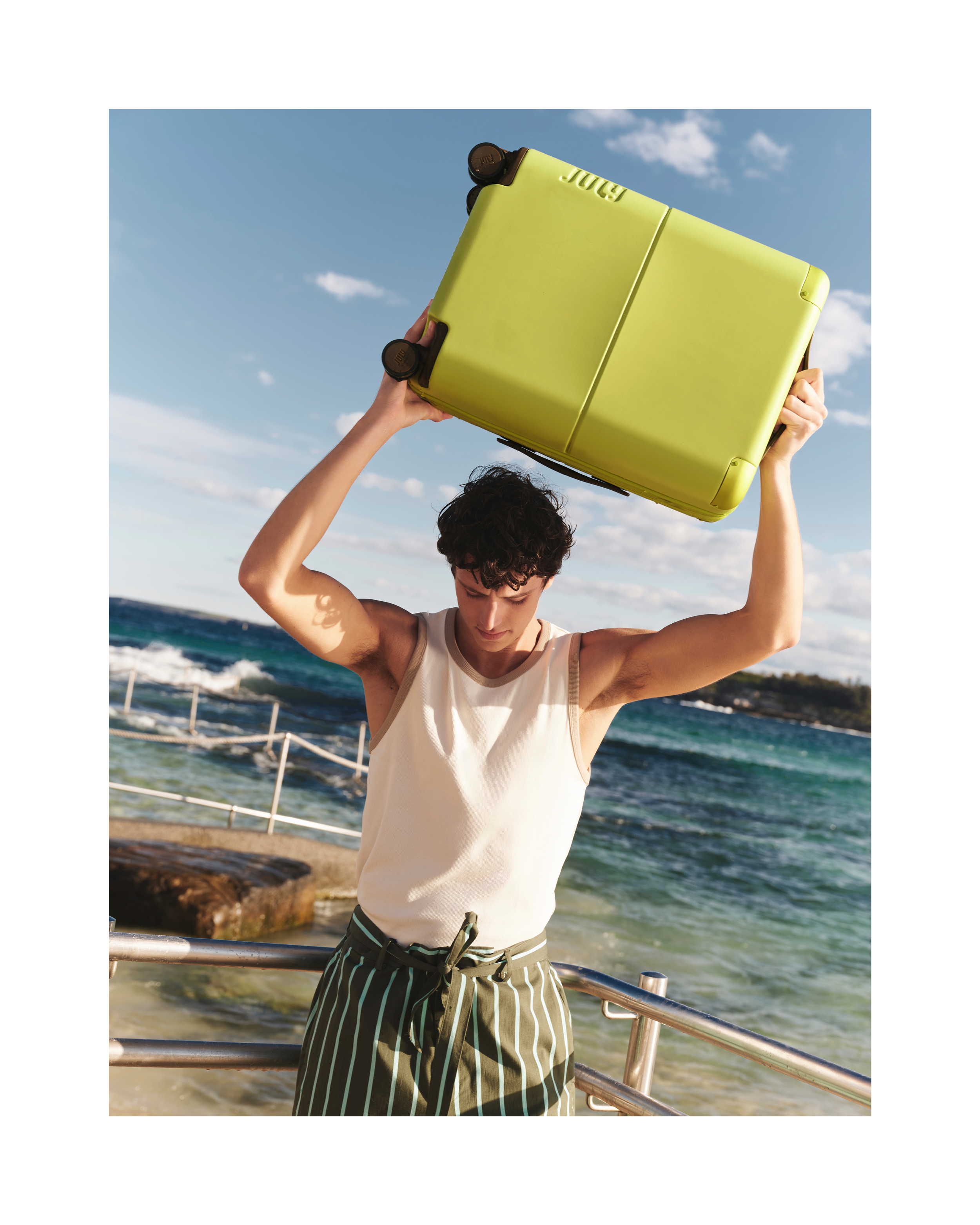 Athletic man in white tank top raises yellow suitcase overhead on ocean coast with blue sky for travel or active lifestyle imagery.