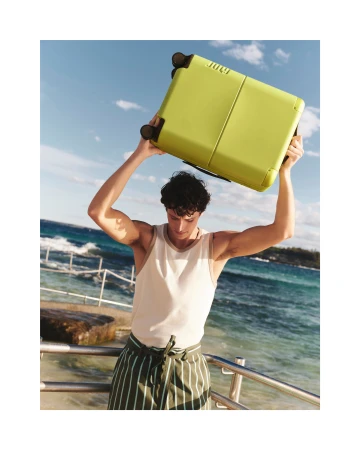 Athletic man in white tank top raises yellow suitcase overhead on ocean coast with blue sky for travel or active lifestyle imagery.