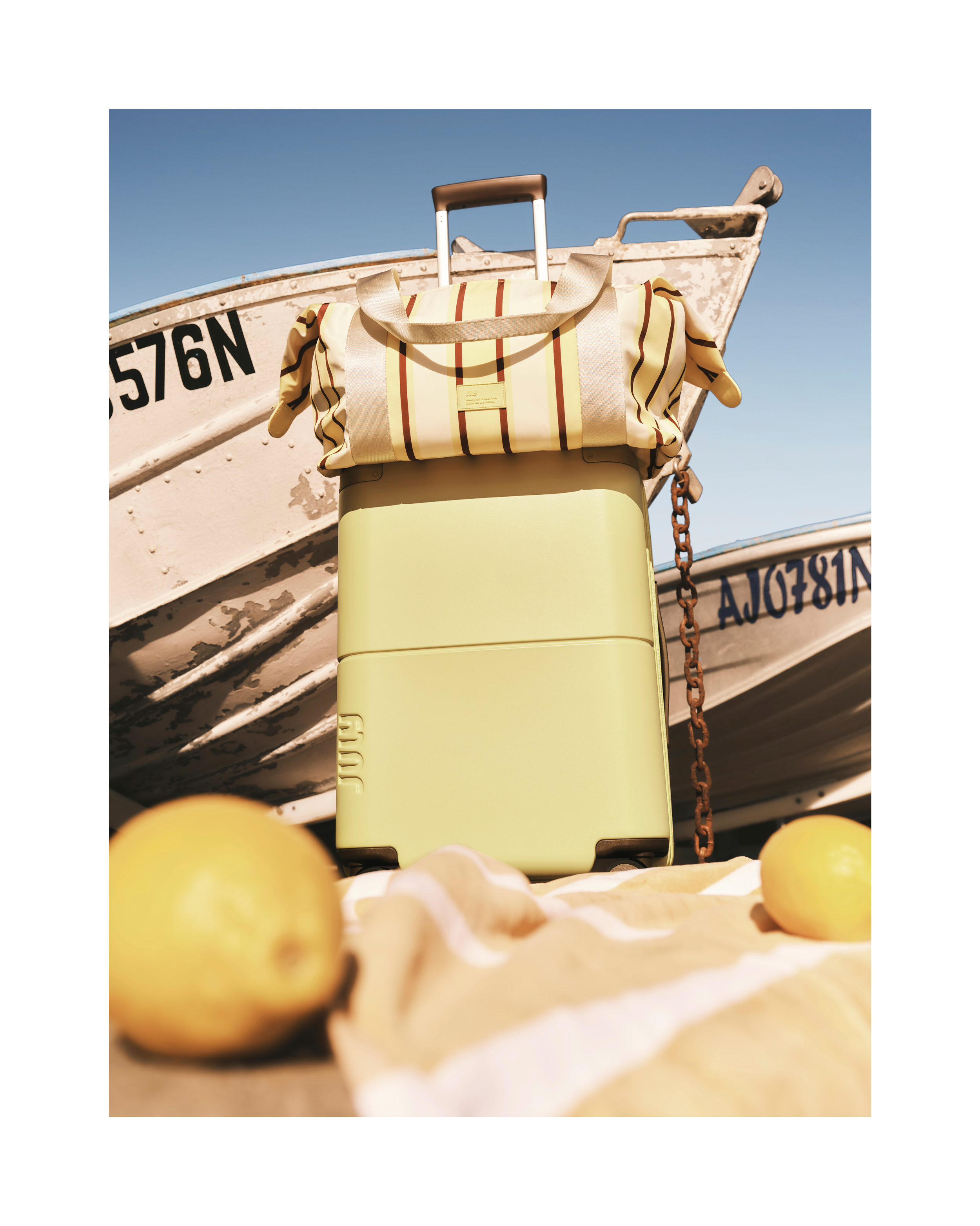 A light green suitcase with a striped bag on top, sits in front of two vintage boats under a blue sky, evoking a travel theme.