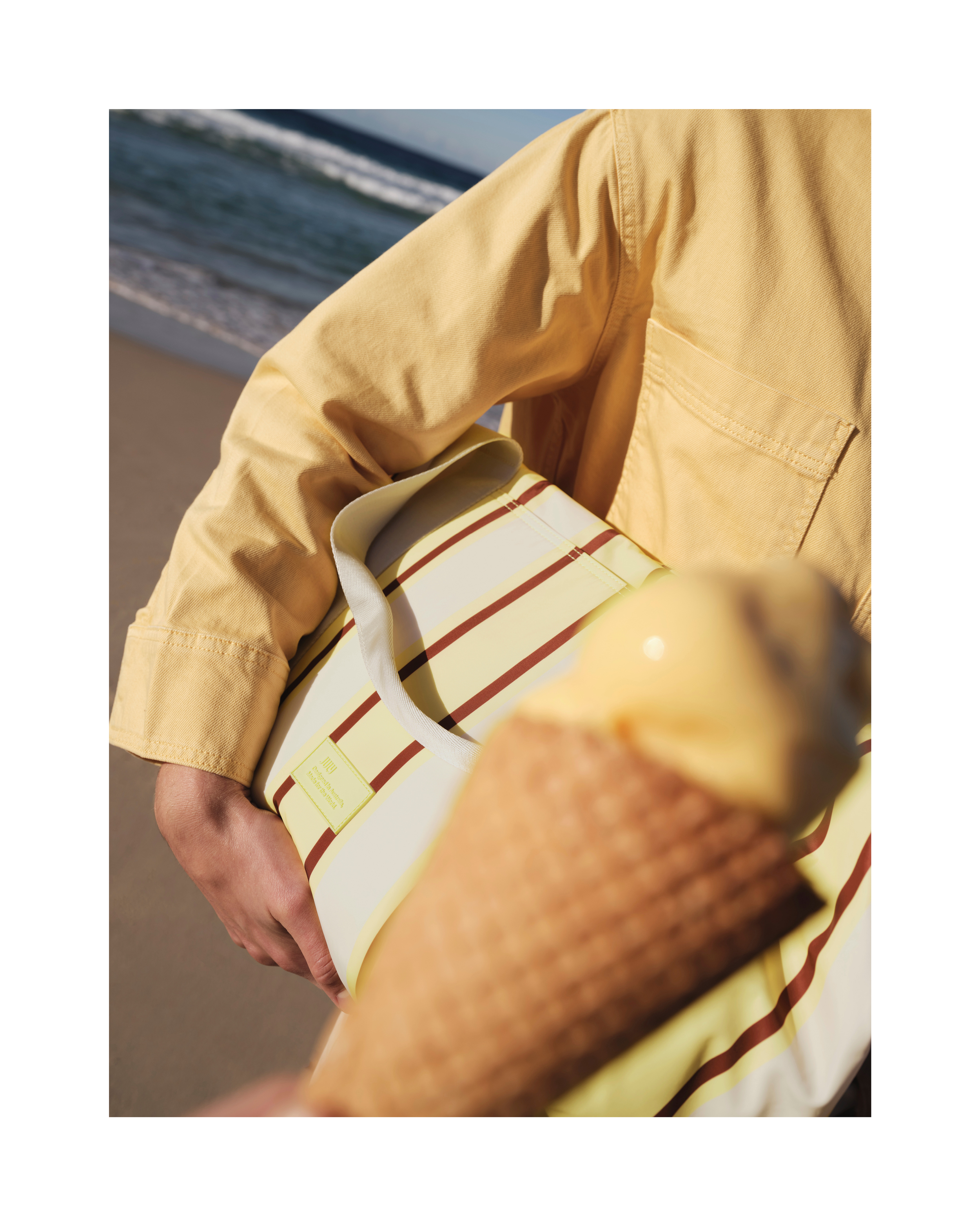 Person in yellow shirt holds striped tote bag with yellow ice cream cone on beach against ocean waves.