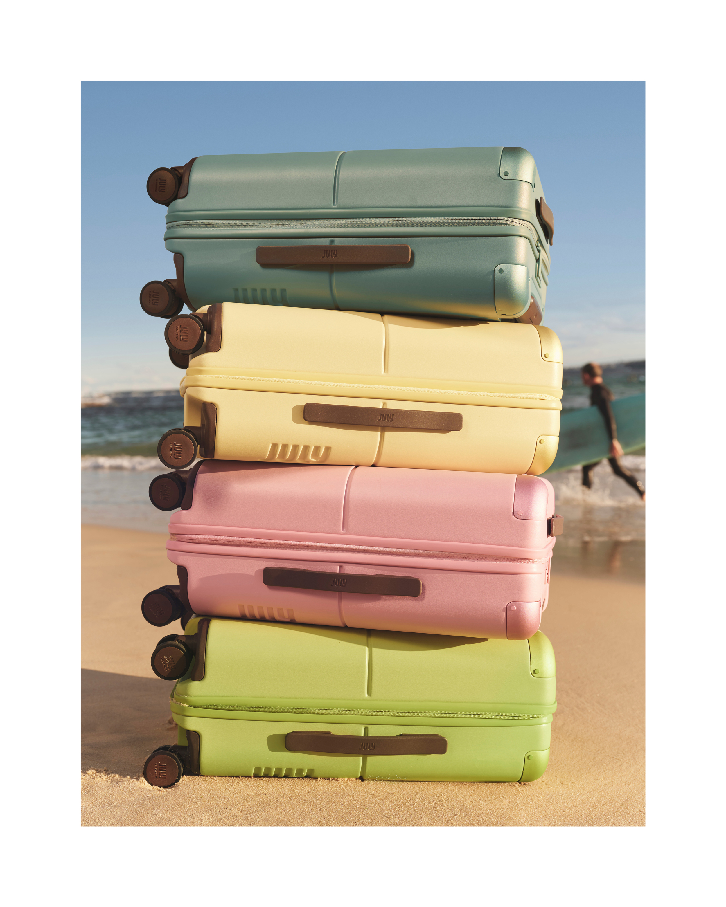 Four pastel hard-shell suitcases stacked vertically on a sandy beach with an ocean backdrop and a distant surfer.