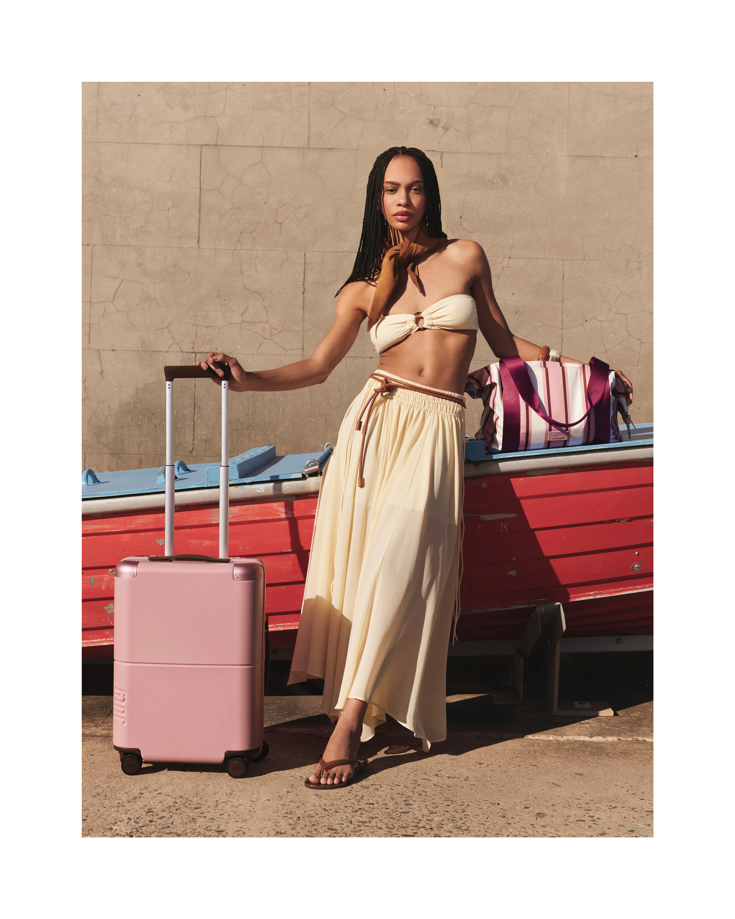 A woman in a bikini top and skirt poses next to a pink suitcase and a striped tote bag in an outdoor setting with natural light.