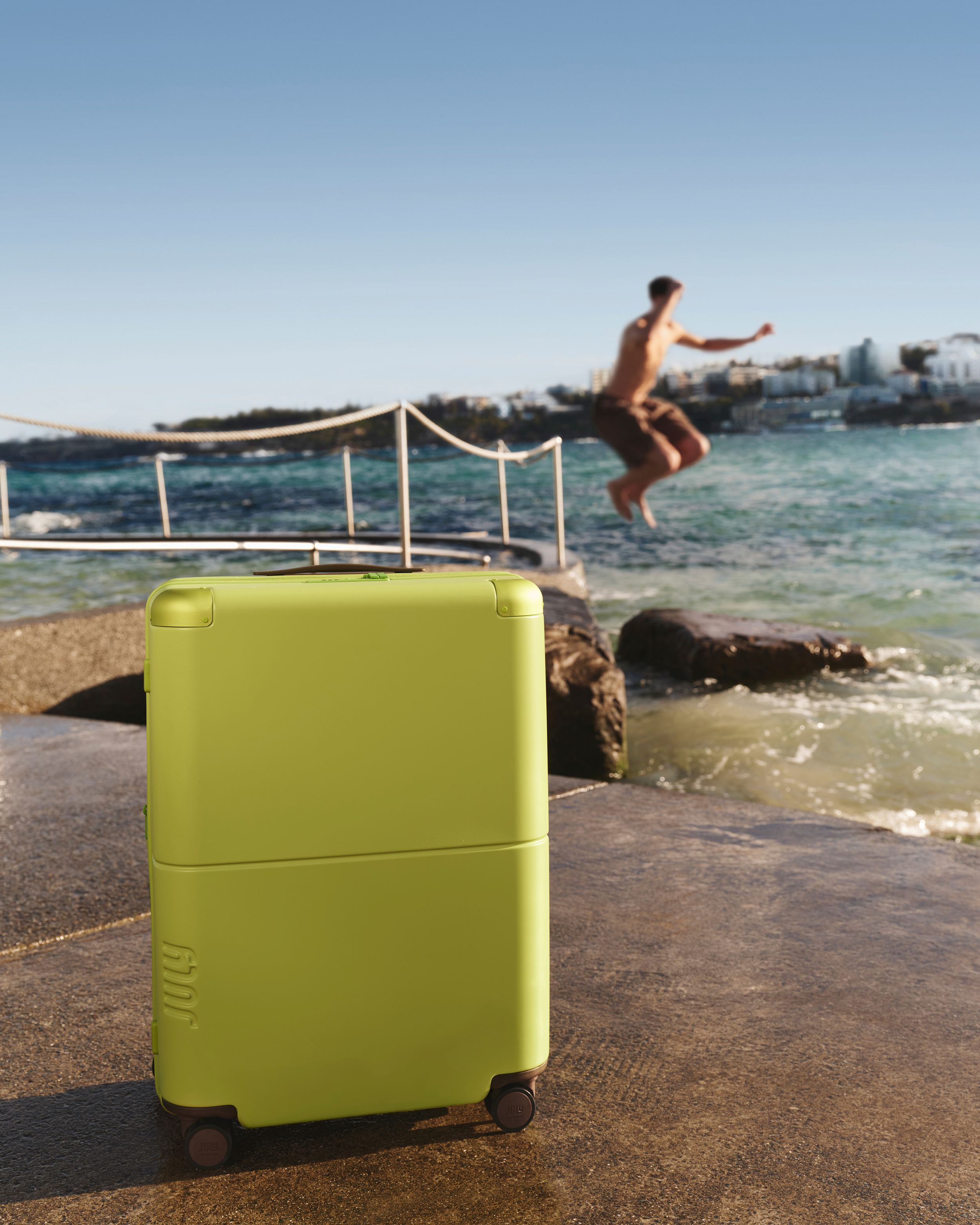 A lime-green suitcase stands by the ocean as a man jumps into the water, with clear skies and coastal architecture in the background.