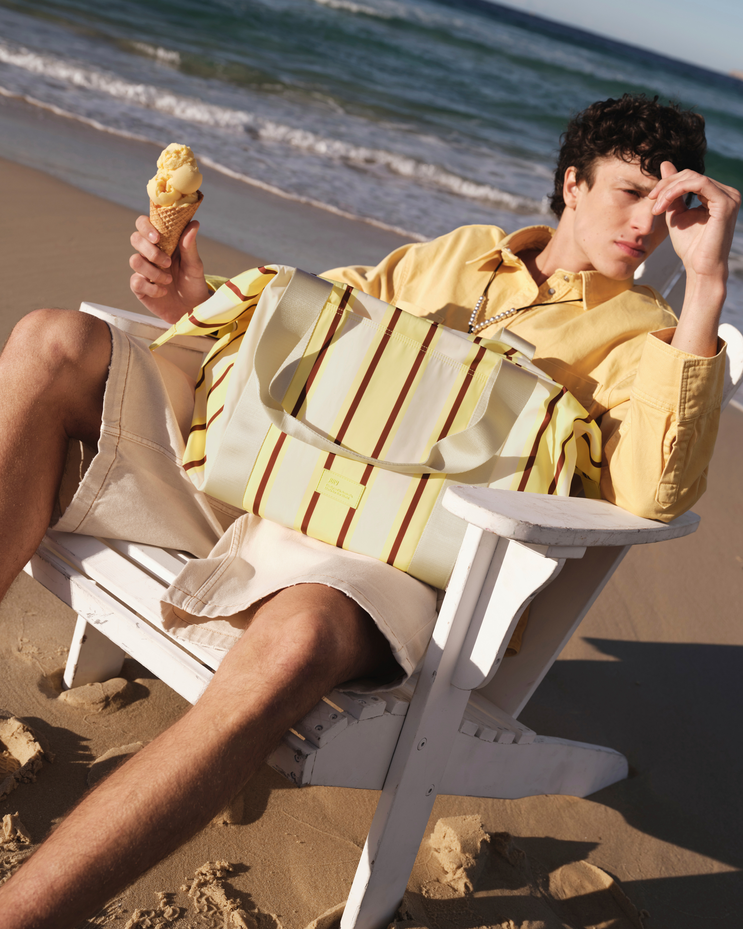 Young adult male relaxes in a white Adirondack chair on a sandy beach, holding an ice cream cone; ocean waves visible in background.