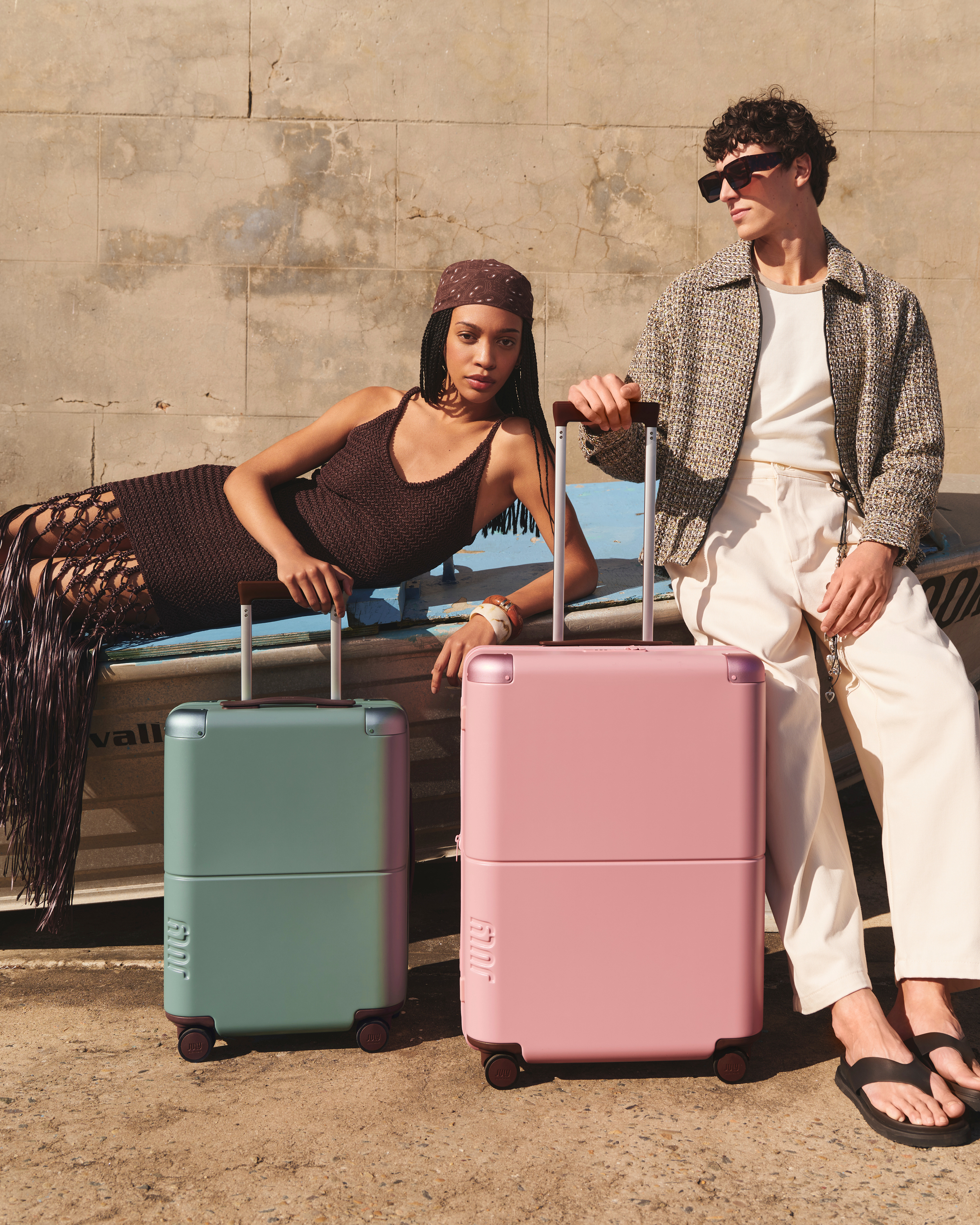 Two travelers pose outdoors with a mint and pink rolling suitcases on a neutral background, showcasing travel and fashion.