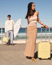 Woman in a striped knit dress standing with yellow luggage on a beach promenade beside a man holding a surfboard.