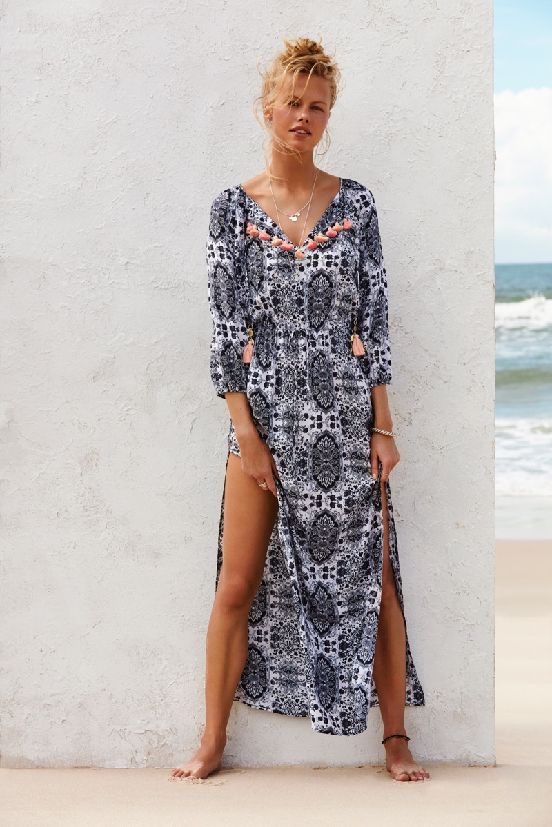 Model standing outdoors wearing a long printed blue and white dress with tassel details against a white wall.