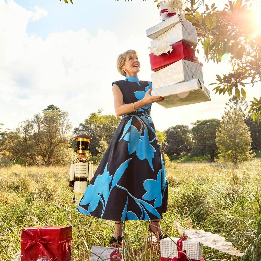 A mature woman stands outside in a field holding a stack of gift-wrapped presents while surrounded by gifts and a toy nutcracker.