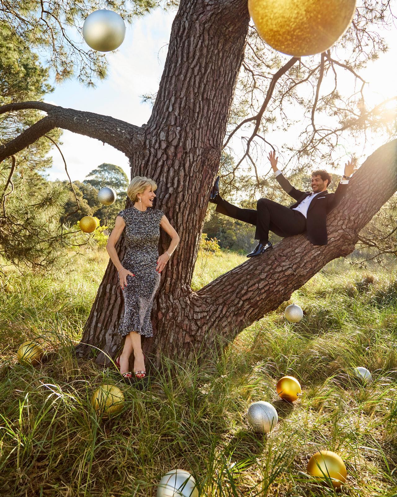 Elegant woman in dress stands by a tree while a man in a suit lounges on a branch, outdoors with gold and silver decorative spheres.