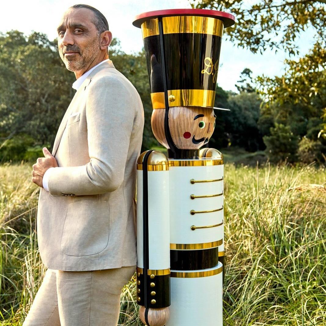 Man in a suit stands in a field next to a large wooden toy soldier, trees are in the background, with soft sunlight filtering through.