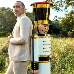 Man wearing a beige suit standing next to a large decorative nutcracker in an outdoor grassy field