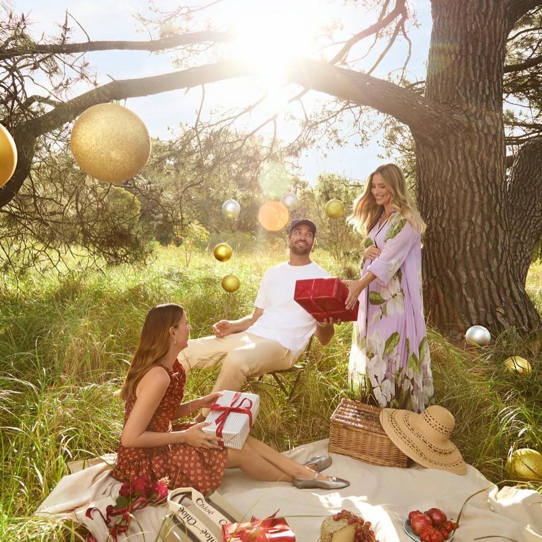Three people exchanging gifts at an outdoor picnic, surrounded by trees and golden ornaments, bathed in soft natural light.