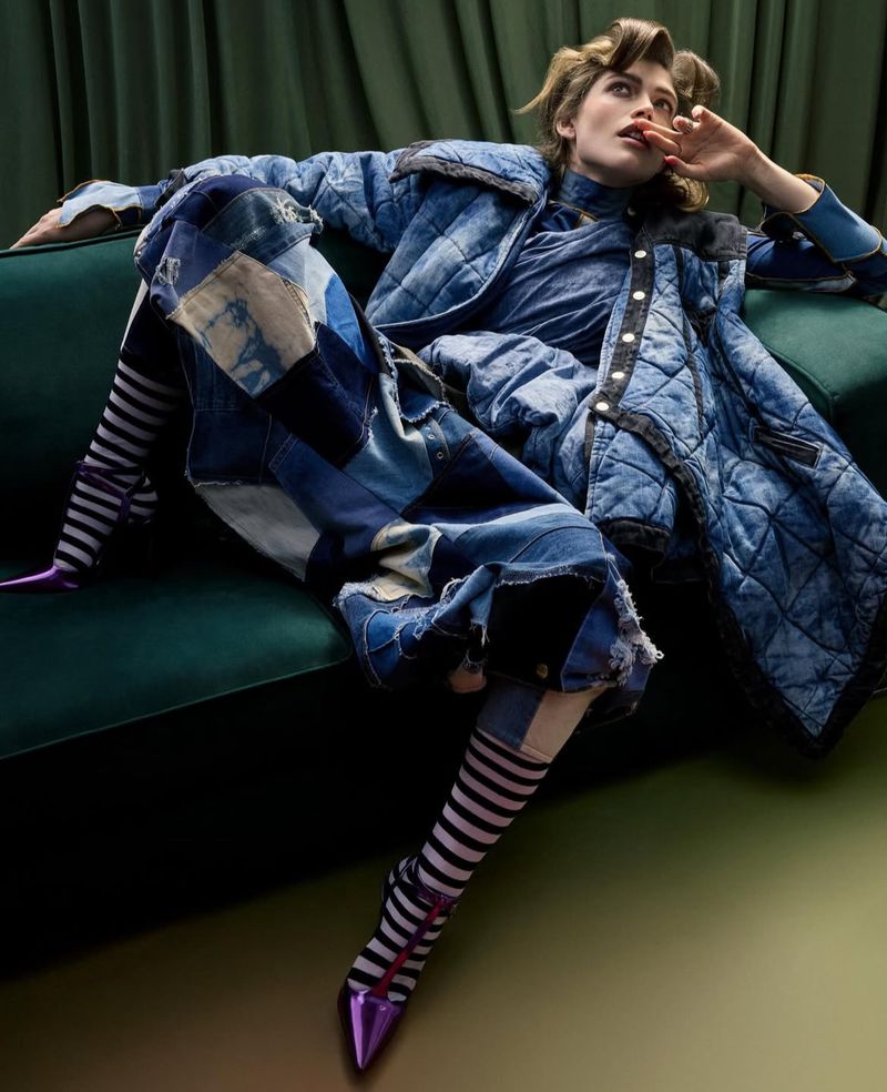 A fashion shot of a woman in a blue quilted jacket and denim patchwork skirt reclining on a green velvet couch, with striped heels and stockings.