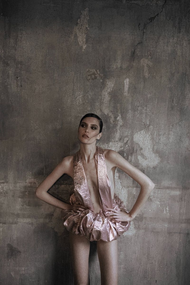 Fashion shot of an adult woman in a metallic pink dress, hands on hips, against a textured grey background in a photo studio setting.