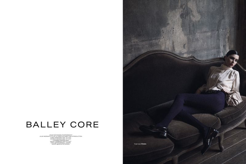 Fashion shot of a woman in a cream blouse and dark pants sitting on a grey couch in a dark interior, photographed in a studio.