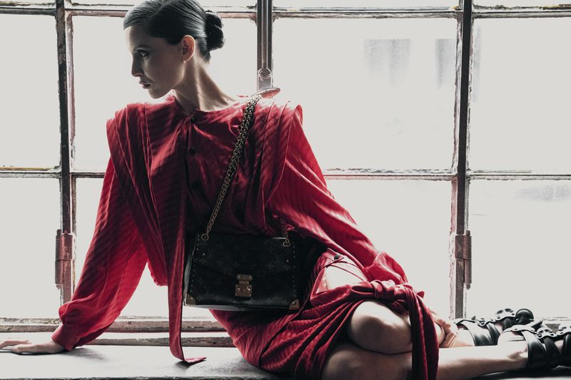 Model in red dress wearing dark shoulder bag seated against a gridded industrial window background