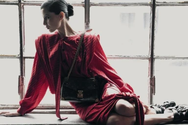 Model in red dress wearing dark shoulder bag seated against a gridded industrial window background