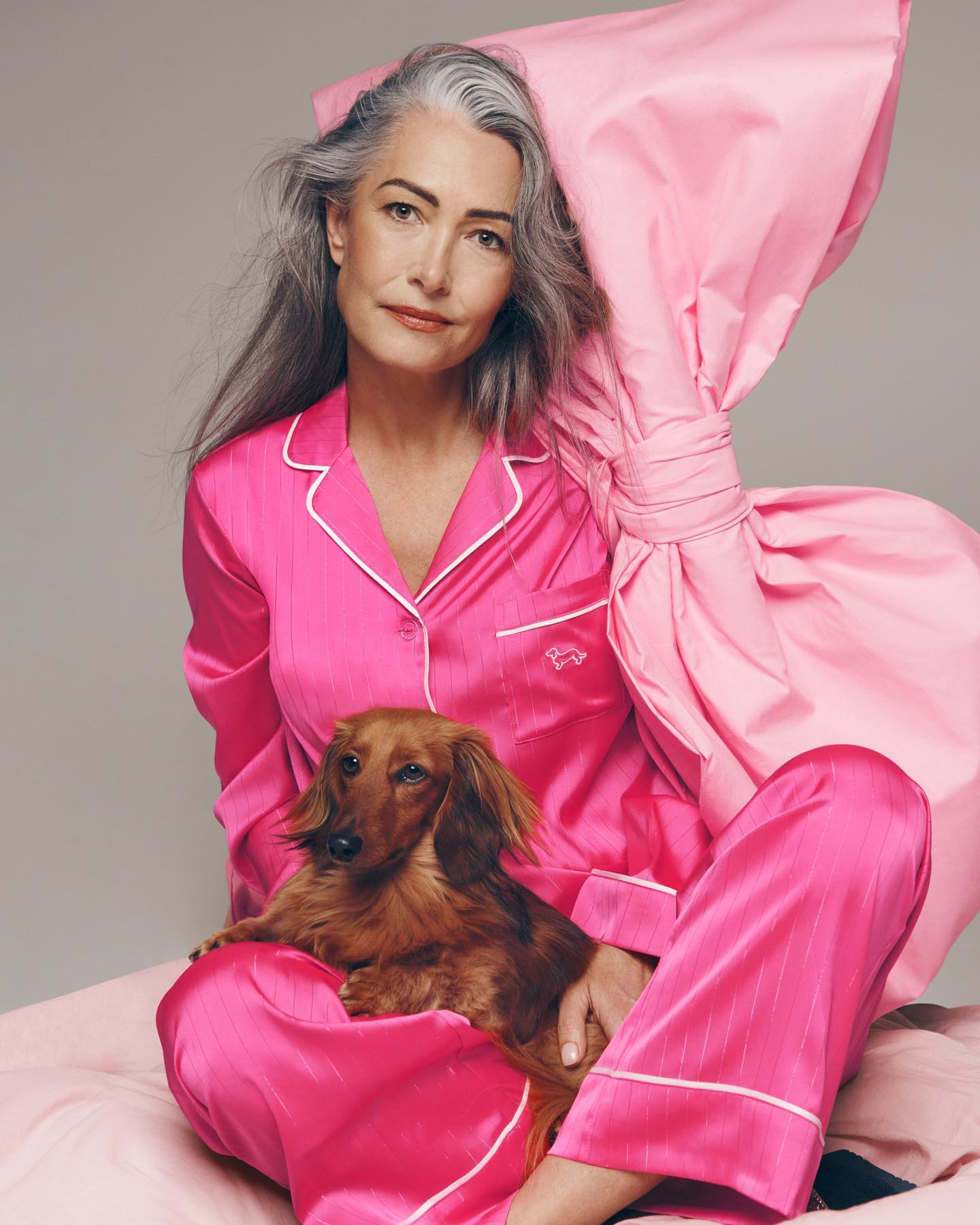 Attractive mature woman with grey hair poses with a dachshund while sitting on a bed with pink satin sheets and a matching pillow.
