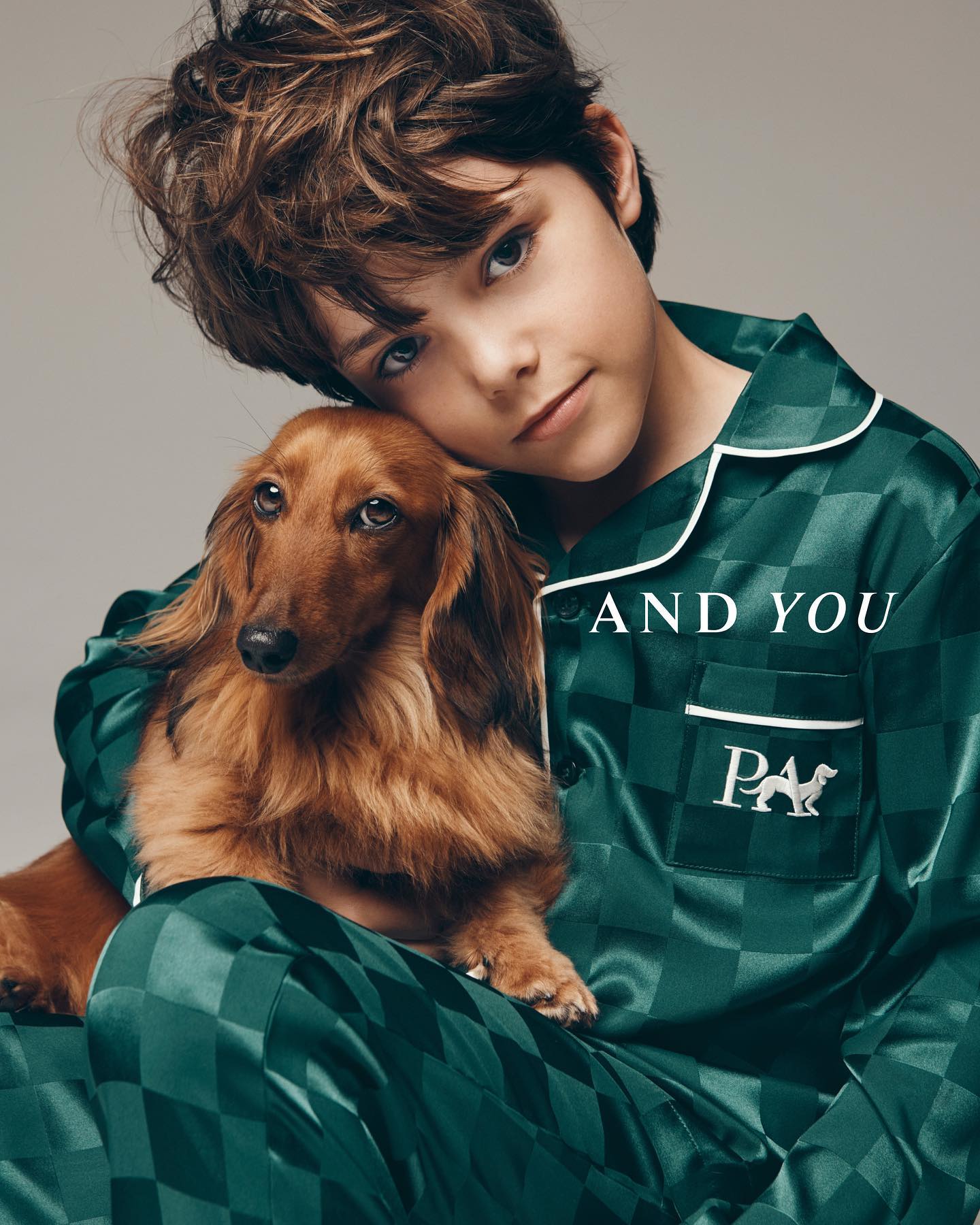 Boy in green checkered pajamas poses with a long-haired dachshund against a grey background. Clothing and pet photography.