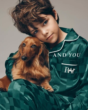 Boy in green checkered pajamas poses with a long-haired dachshund against a grey background. Clothing and pet photography.