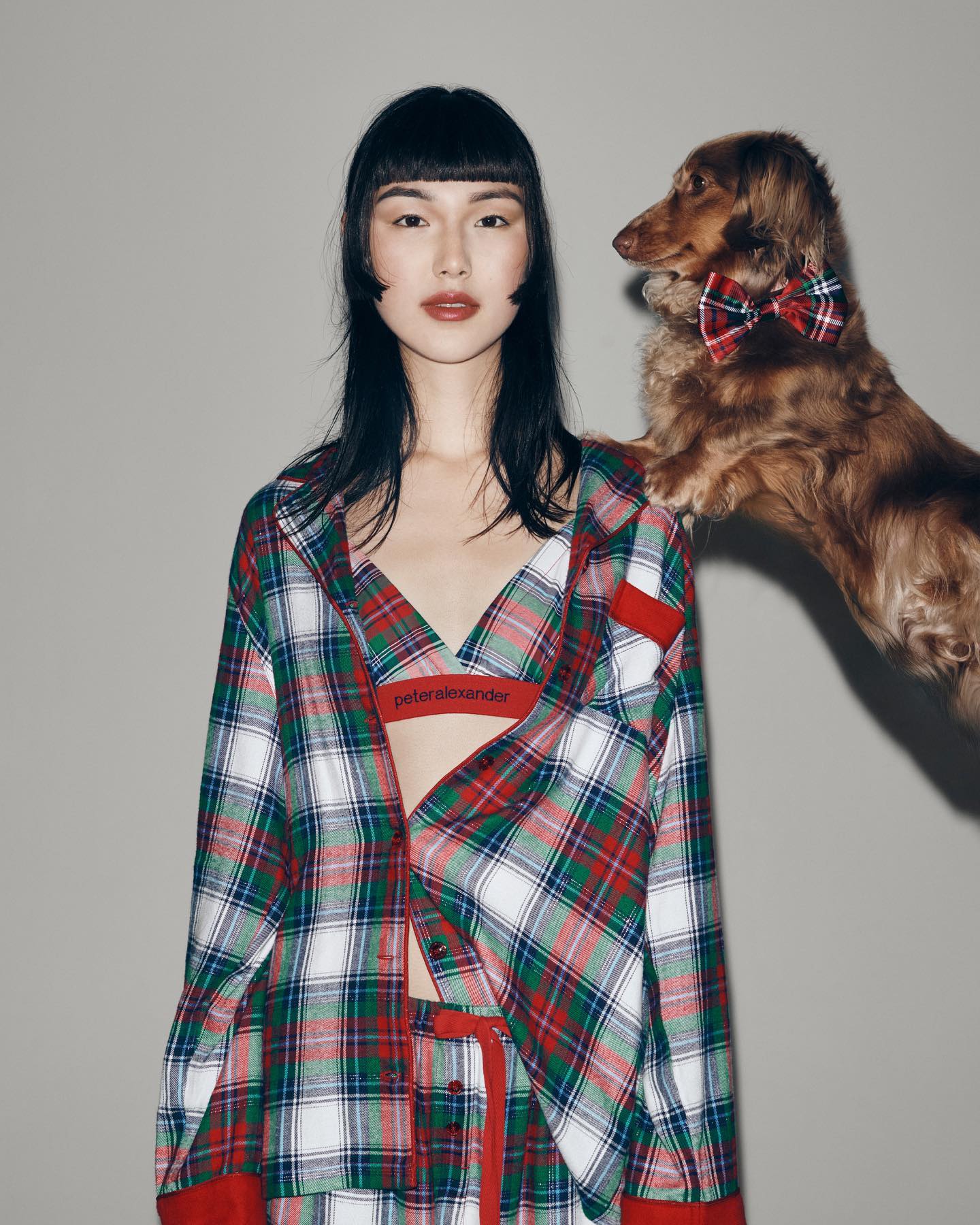 A woman and a dachshund in a photo studio. The woman wears a plaid pajama set and the dog is wearing a matching bow tie.