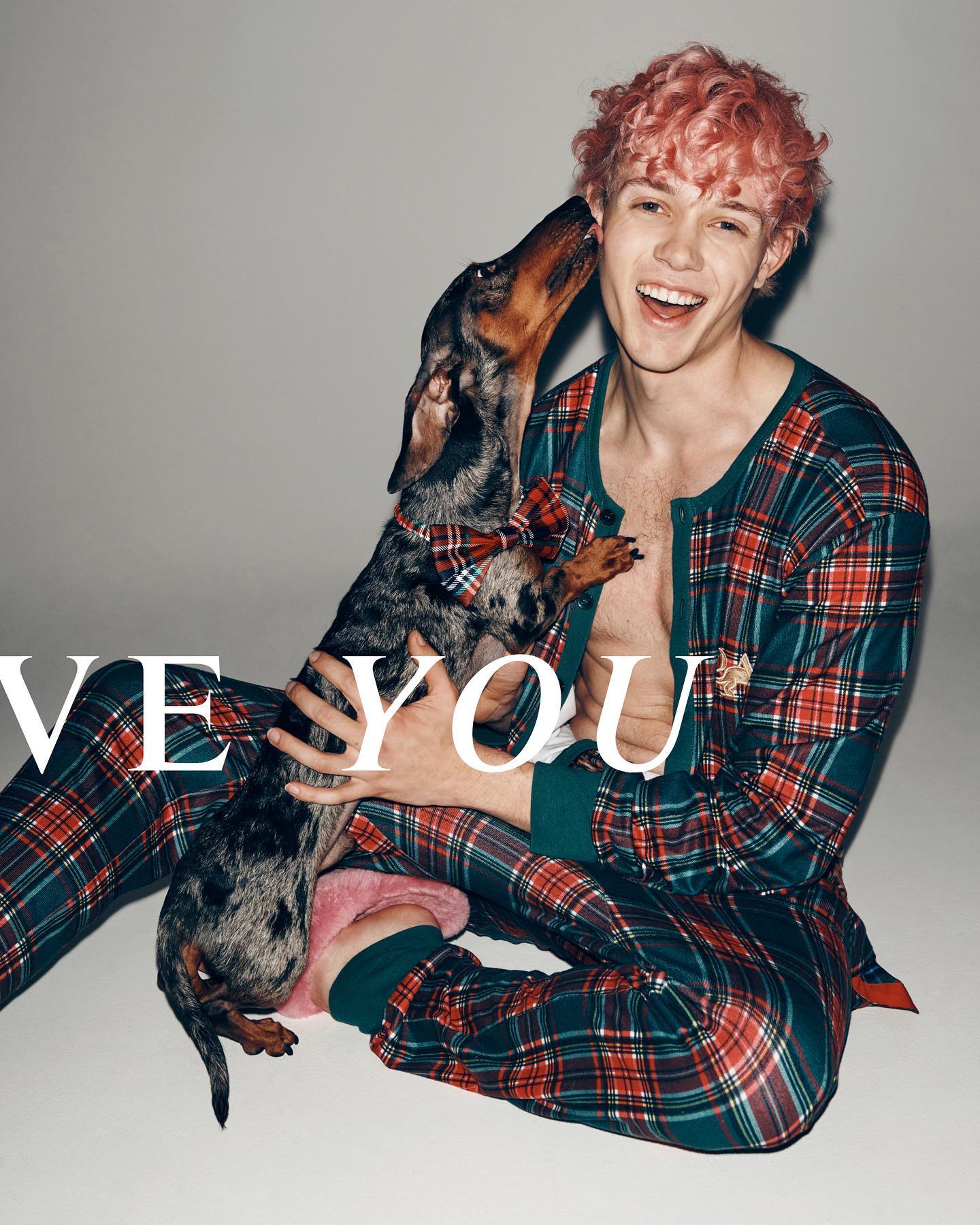 A young adult man with pink hair smiles while holding a dachshund wearing a bow tie in a studio setting with neutral background.