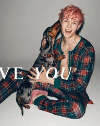 Young man with pink curly hair wearing plaid pyjamas holding a dapple dachshund on a grey studio background.