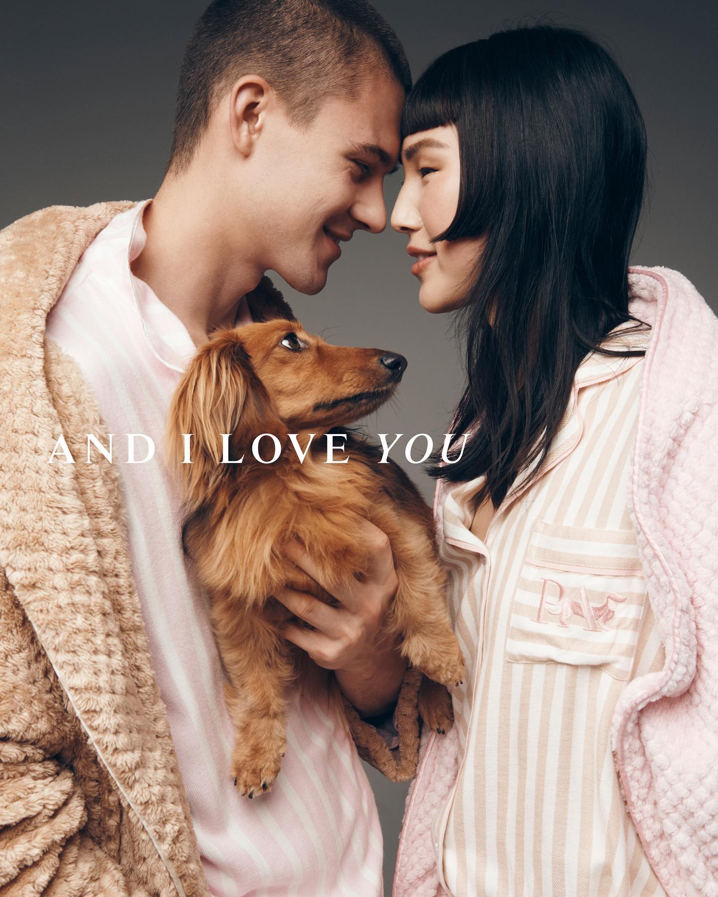 Couple in pajamas poses indoors with their dachshund. The man is wearing a robe and smiling, while the woman's hair is dark and sleek.