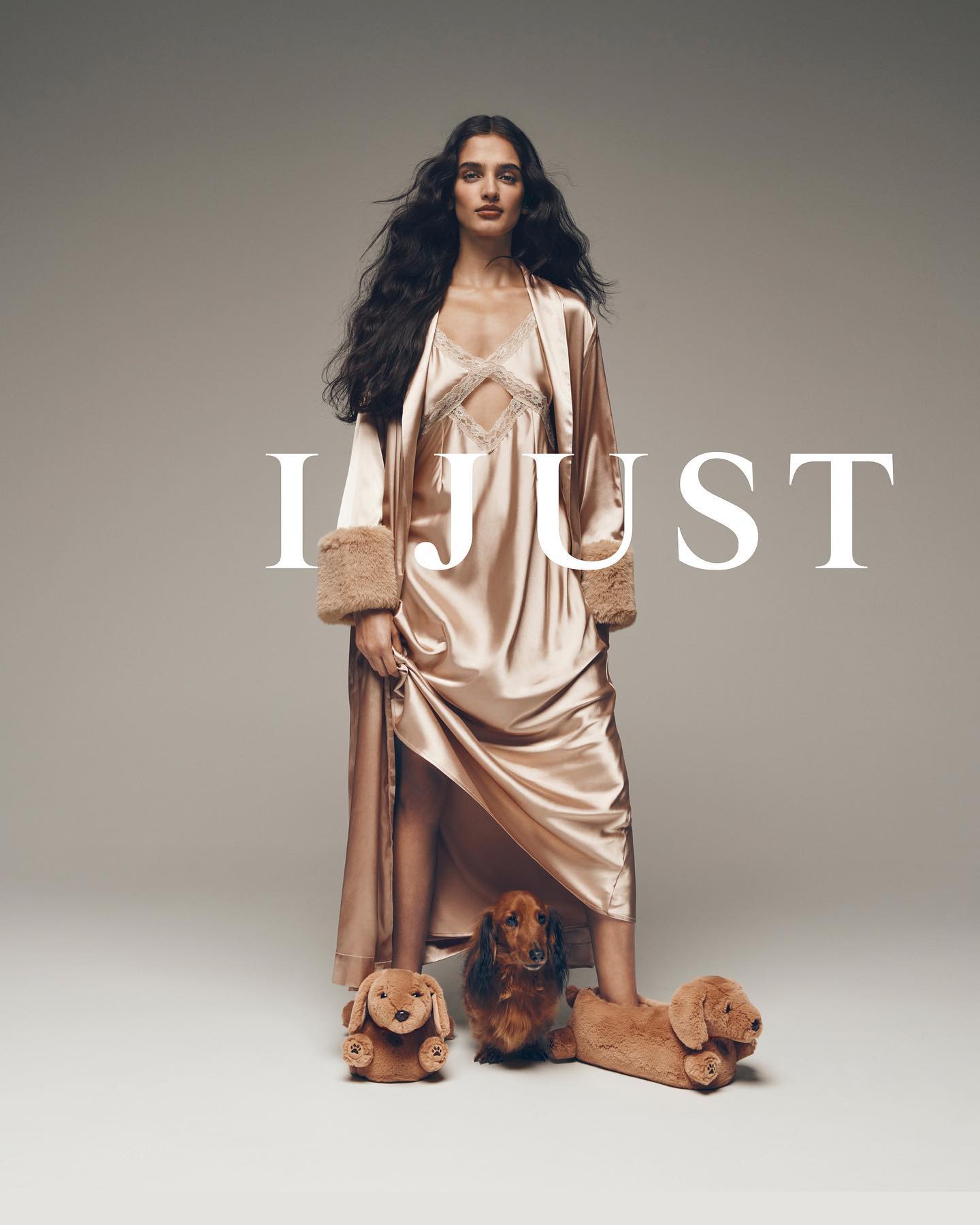 A woman in a gold nightgown and robe poses with dachshund plush toys, standing against a neutral backdrop in a fashion studio.