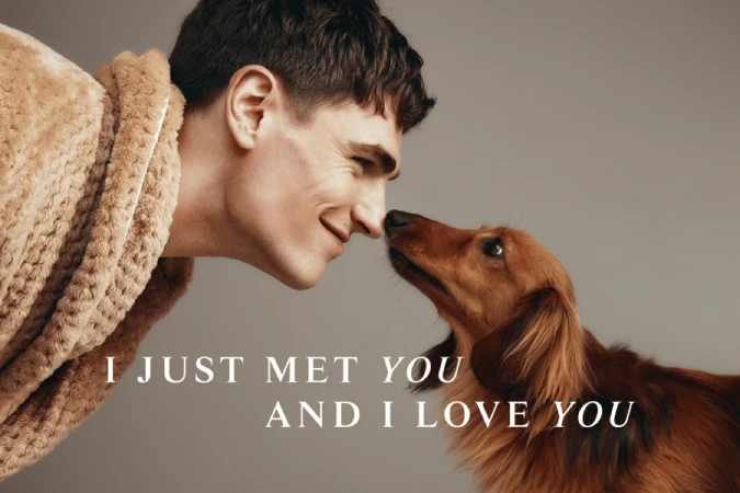 Close-up of a smiling man in a robe touching noses with a long-haired dachshund dog against a neutral grey studio backdrop.