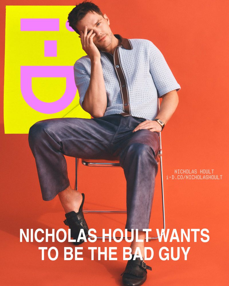 Nicholas Hoult sits on a chair in front of a bright orange background with a magazine cover featuring a yellow rectangle with pink letters.