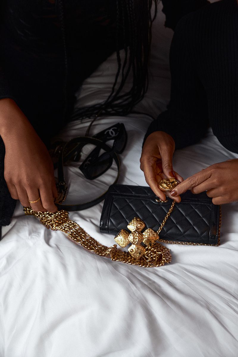Two dark-skinned adults arrange a gold Chanel chain over a black quilted Chanel handbag and sunglasses on a white bedsheet.