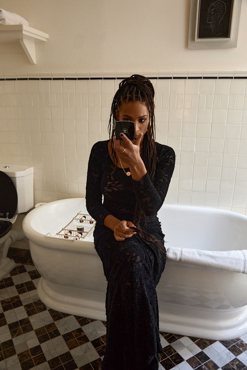 A woman in a black lace dress takes a selfie in a bathroom with white tiles and a freestanding bathtub, creating an elegant self-portrait.