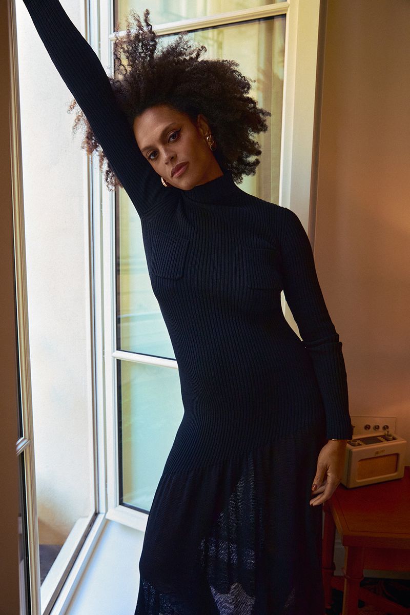 Portrait of a woman in a black ribbed dress posing indoors near a window with soft lighting, showcasing fashion and style.