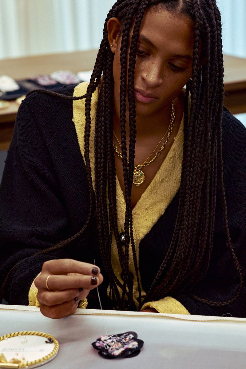 Adult female with braided hair embroiders a flower with needle and thread, wearing a gold necklace and a black and yellow sweater.
