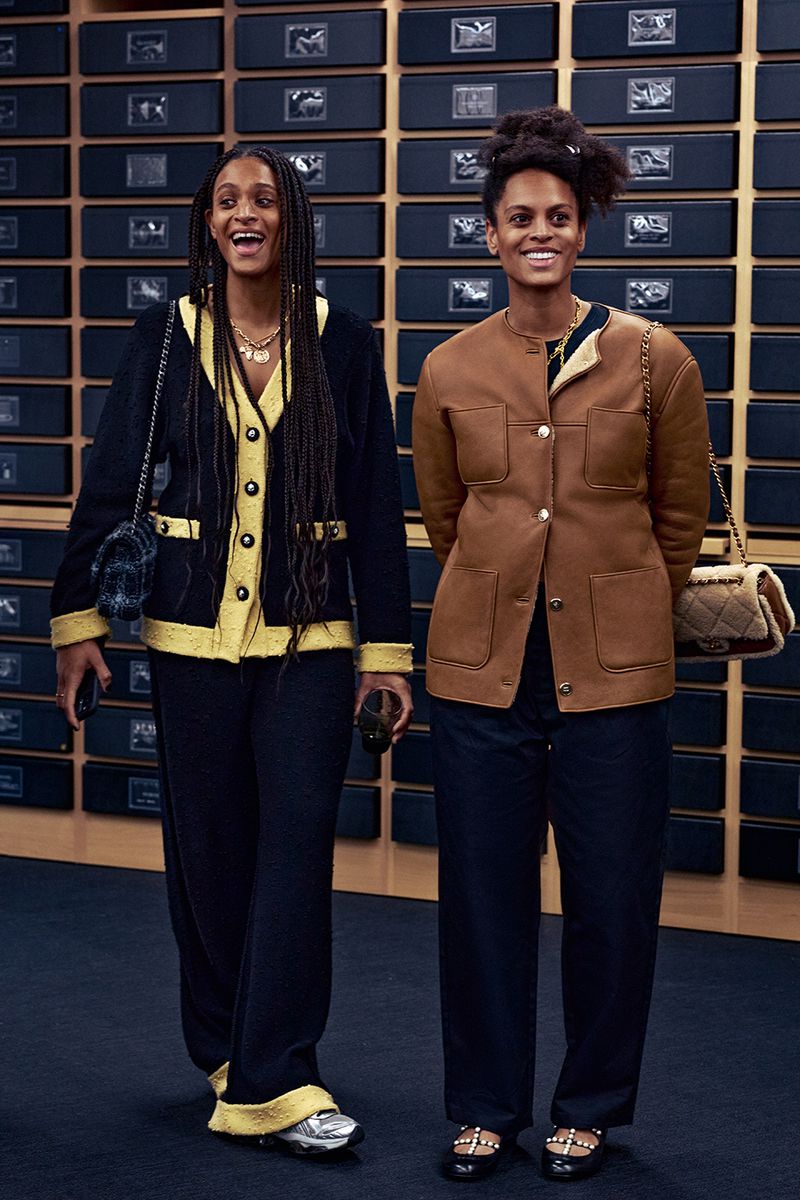 Two women in stylish jackets and dark pants pose in front of a wall with rows of mounted black boxes, indoor, medium shot.