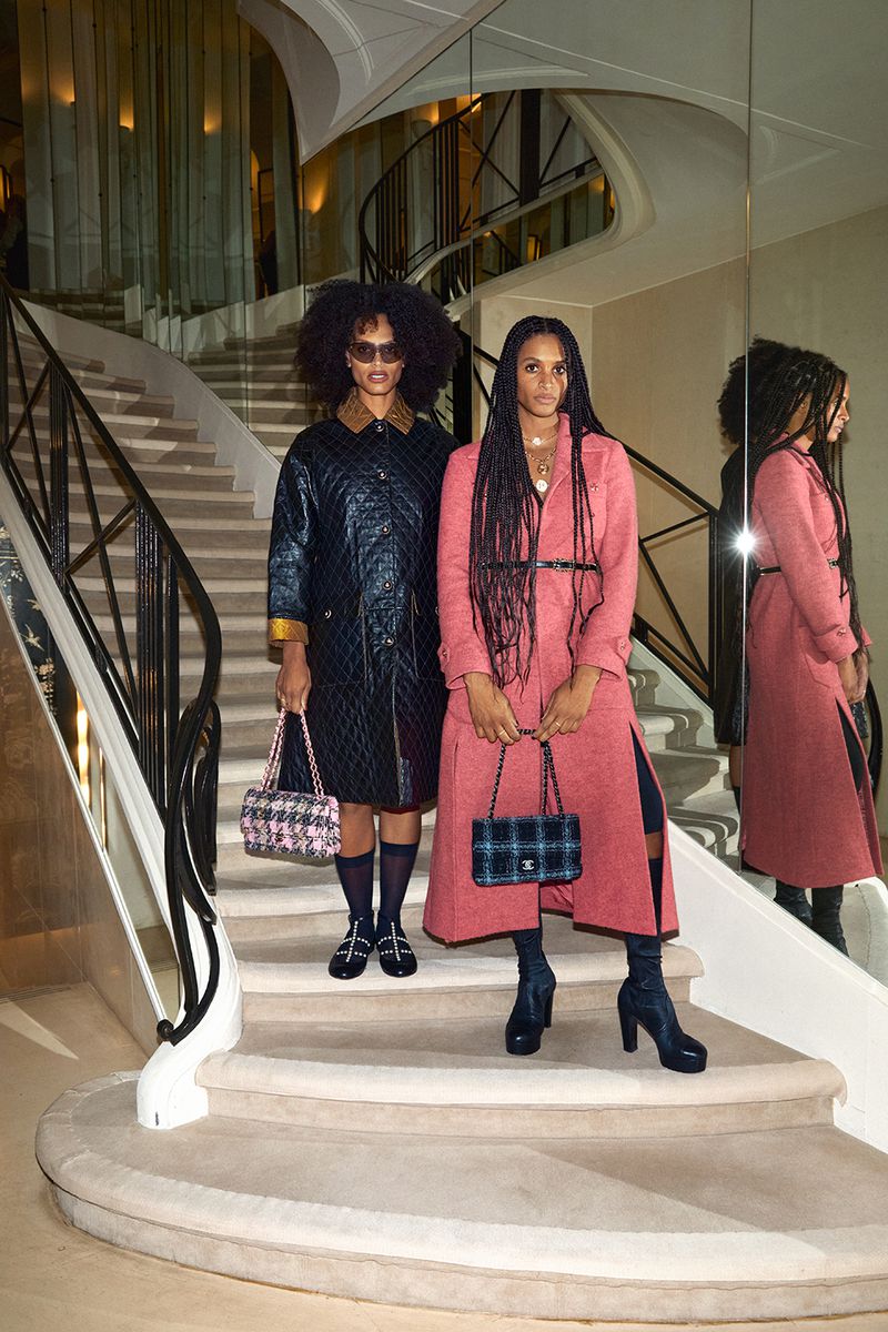 Two women pose on a staircase in fashionable coats and handbags, showcasing modern style with a hint of vintage and high-end design.