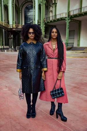 Two fashion models pose in an indoor location wearing winter coats, platform boots, and carrying purses in a medium studio shot.