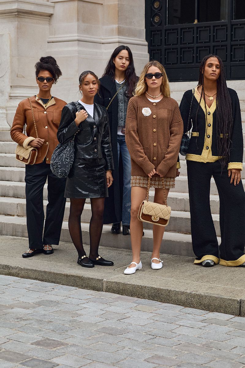 Five women pose on concrete steps wearing fashionable clothing and accessories. Some hold purses and wear sunglasses in an outdoor setting.