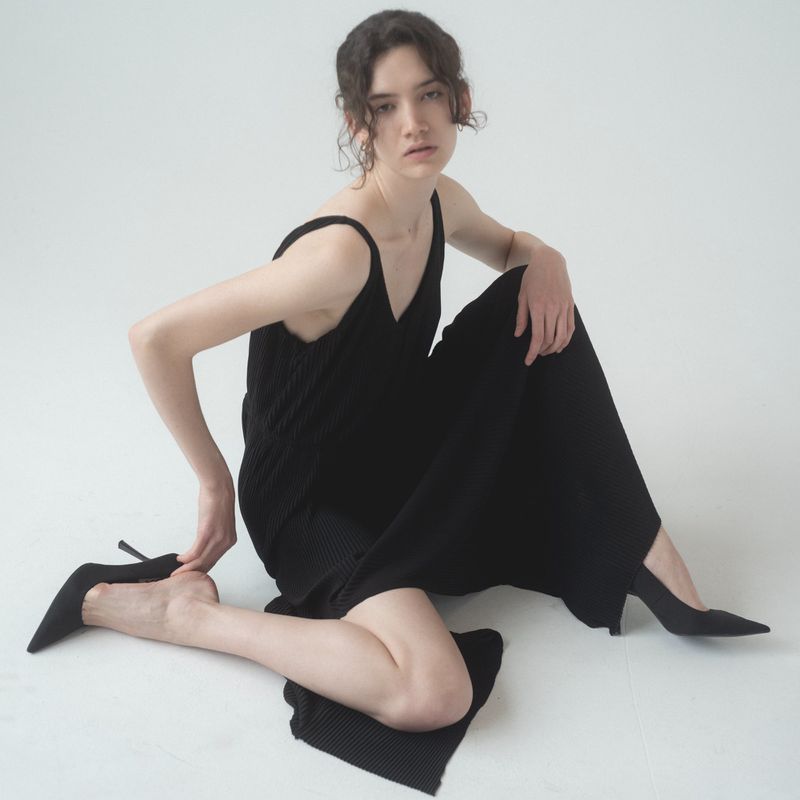 Elegant woman seated on a white backdrop in a black knit dress and heels, striking a pose with soft studio lighting.