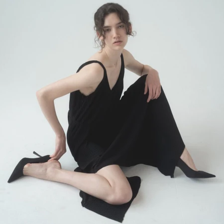 Elegant woman seated on a white backdrop in a black knit dress and heels, striking a pose with soft studio lighting.