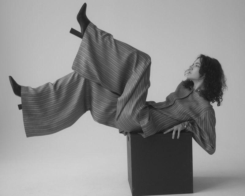 An adult woman in a striped outfit and black heeled boots is reclining on a dark cube in a studio with a bright background.