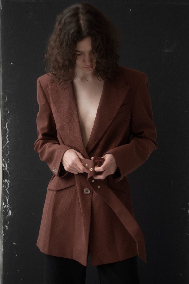 A person with curly hair wears a brown blazer with a belt, standing in front of a black wall. The blazer is buttoned at the waist.