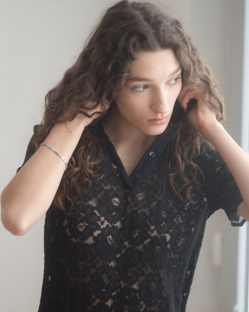 Portrait of a woman with long curly brown hair wearing a black lace top and a bracelet, posing indoors in front of a neutral wall.