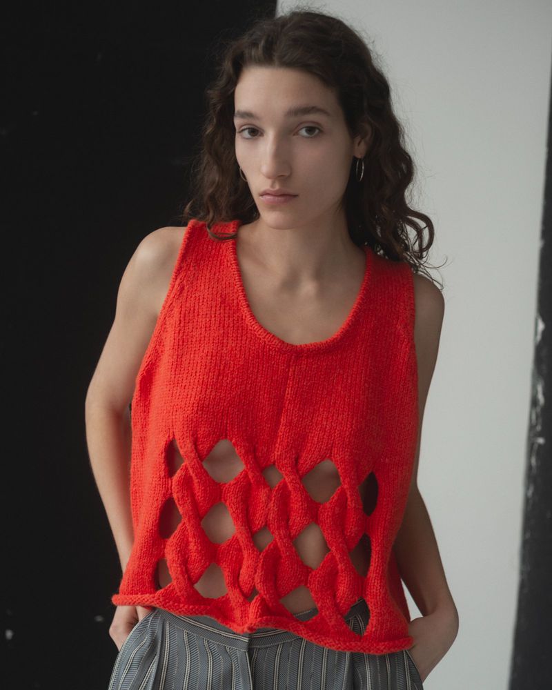 Young woman with long wavy brown hair poses indoors wearing a bright red knitted top with a geometric cutout pattern in front of black and white panels.