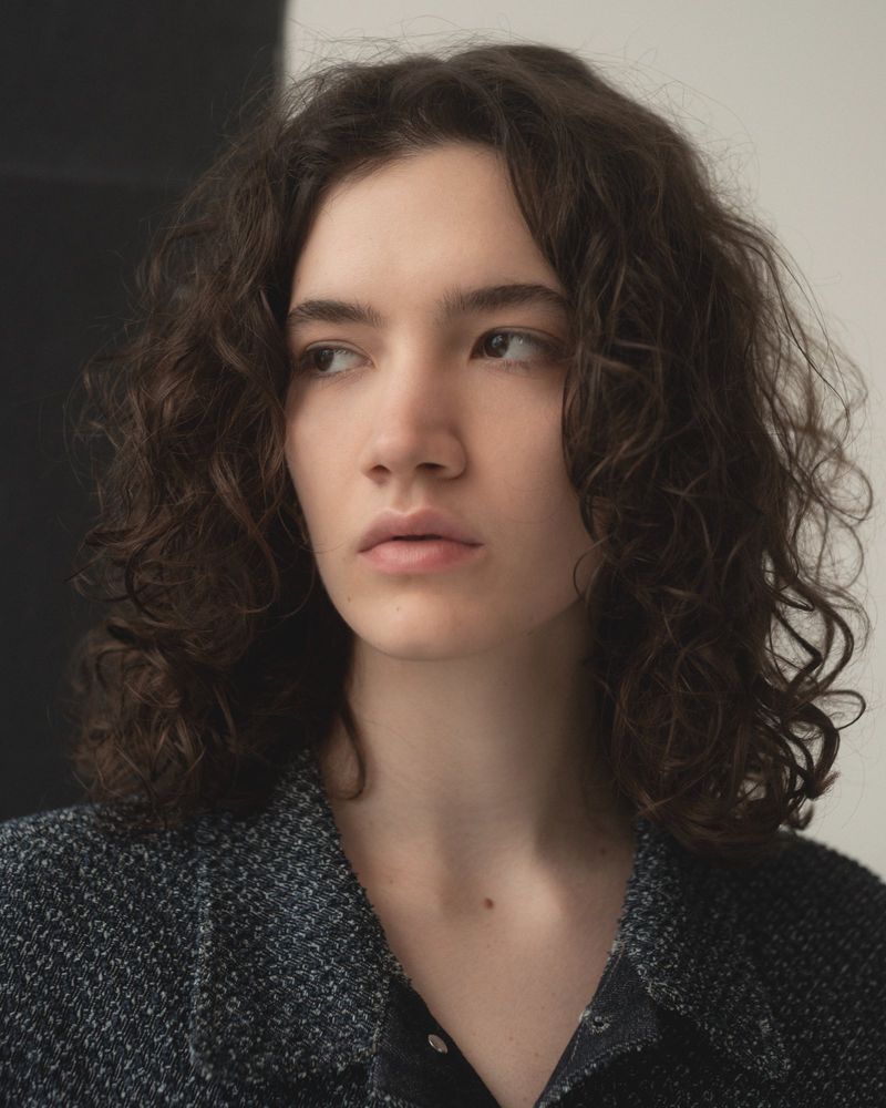 Portrait of a young woman with curly brown hair wearing a dark tweed jacket against a neutral studio background