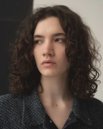 Portrait of a young woman with curly brown hair wearing a dark tweed jacket against a neutral studio background