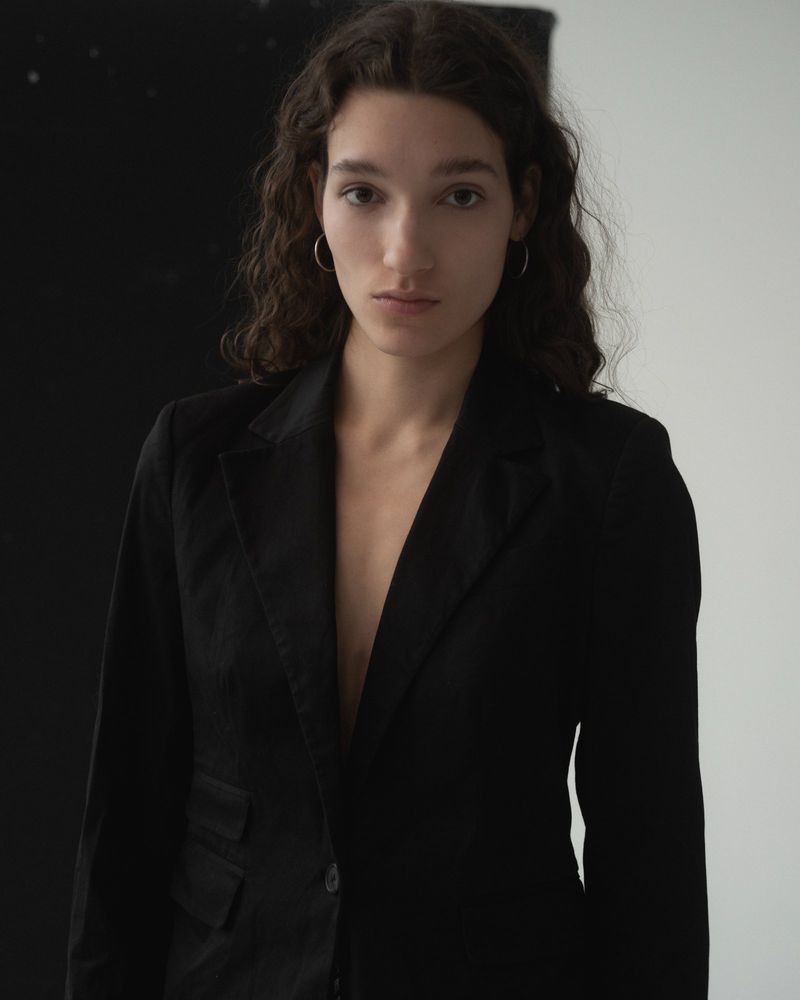 Portrait of a woman with dark wavy hair wearing a black blazer against a split black and white background.